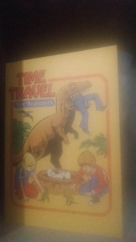 A birthday card captioned "Time travel for beginners". A retro style cartoon shows two children playing with dinosaur hatchlings while a large theropod dinosaur looms behind them, human legs protruding from its mouth 