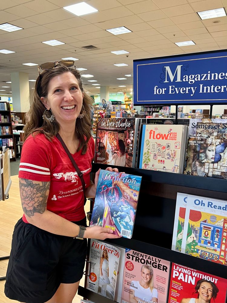A woman with shoulder length hair, wearing red dinosaur t-shirt and sunglasses on her head smiling while holding a copy of an illustrated magazine in a bookstore.