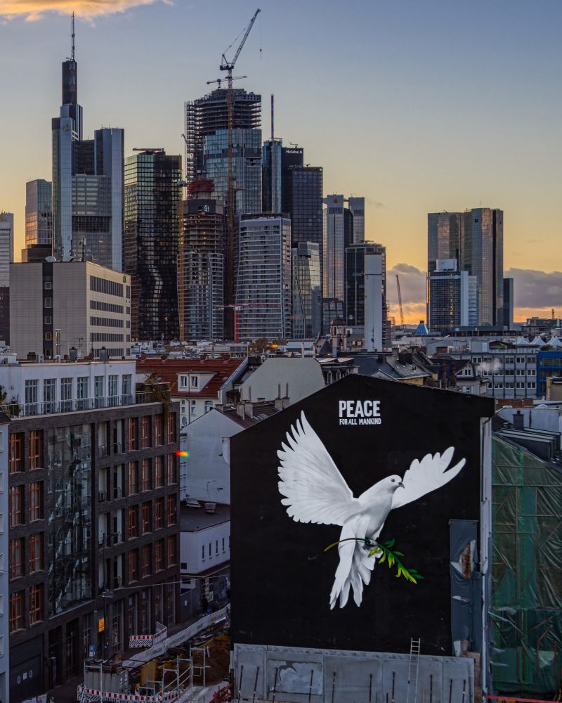 „PEACE FOR ALL MANKIND“ steht über dem Bild einer riesigen Friedenstaube an einer Hauswand in Frankfurt am Main, am Horizont die städtische Skyline, © picture alliance / greatif, Foto: Florian Gaul