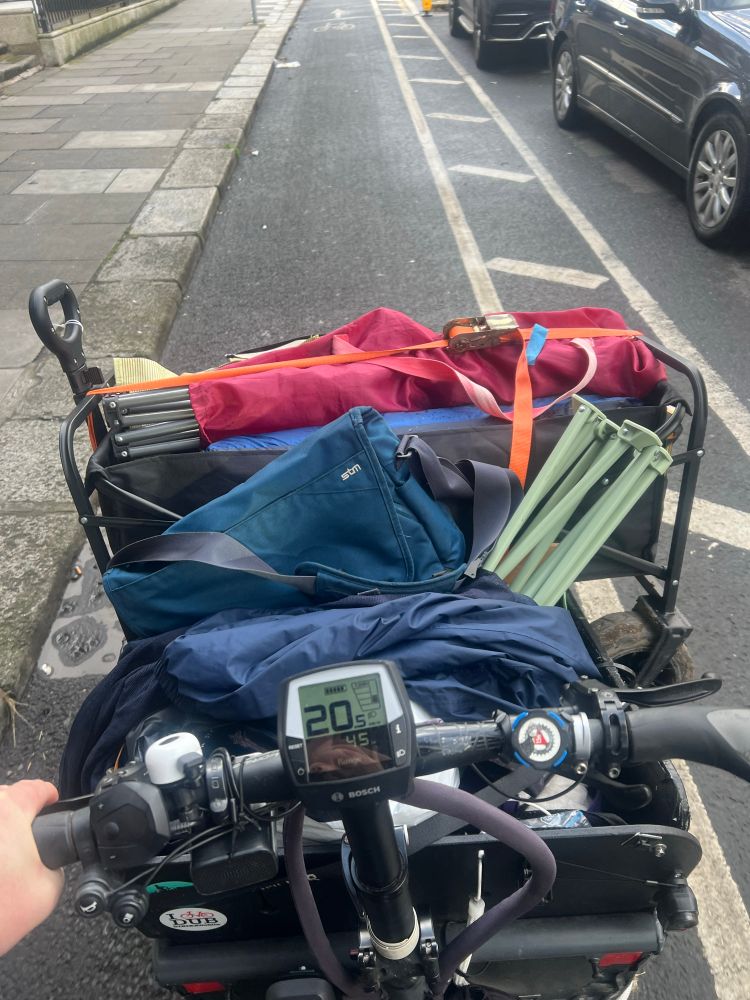 Cargo bike loaded with various camping gear including a cart, chairs, tent sleeping bag. POV from behind the handlebars 