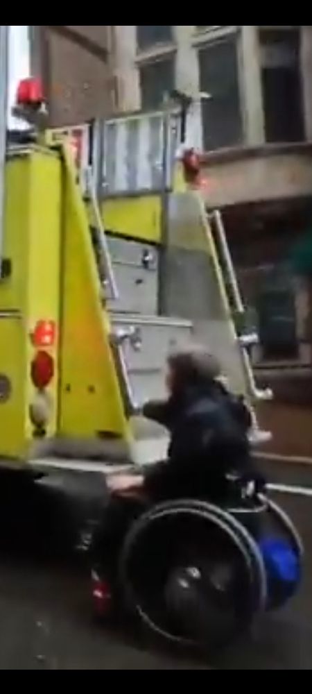 Protester in a manual wheelchair catches a ride on the back of a firetruck to avoid pushing up the hill