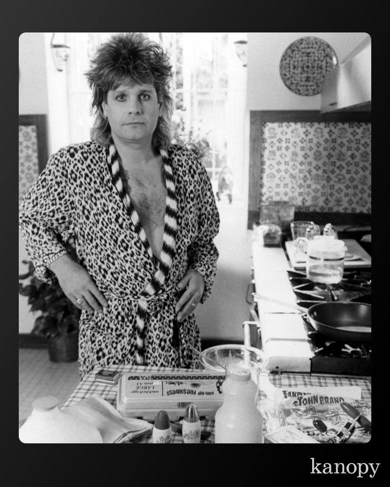 Black and white image of Ozzy Osbourne standing in a kitchen while wearing a leopard print bathrobe.
