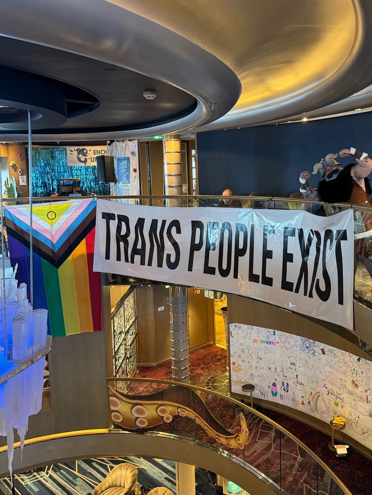A large banner with the inclusive Pride flag and a sign that says “trans people exist”