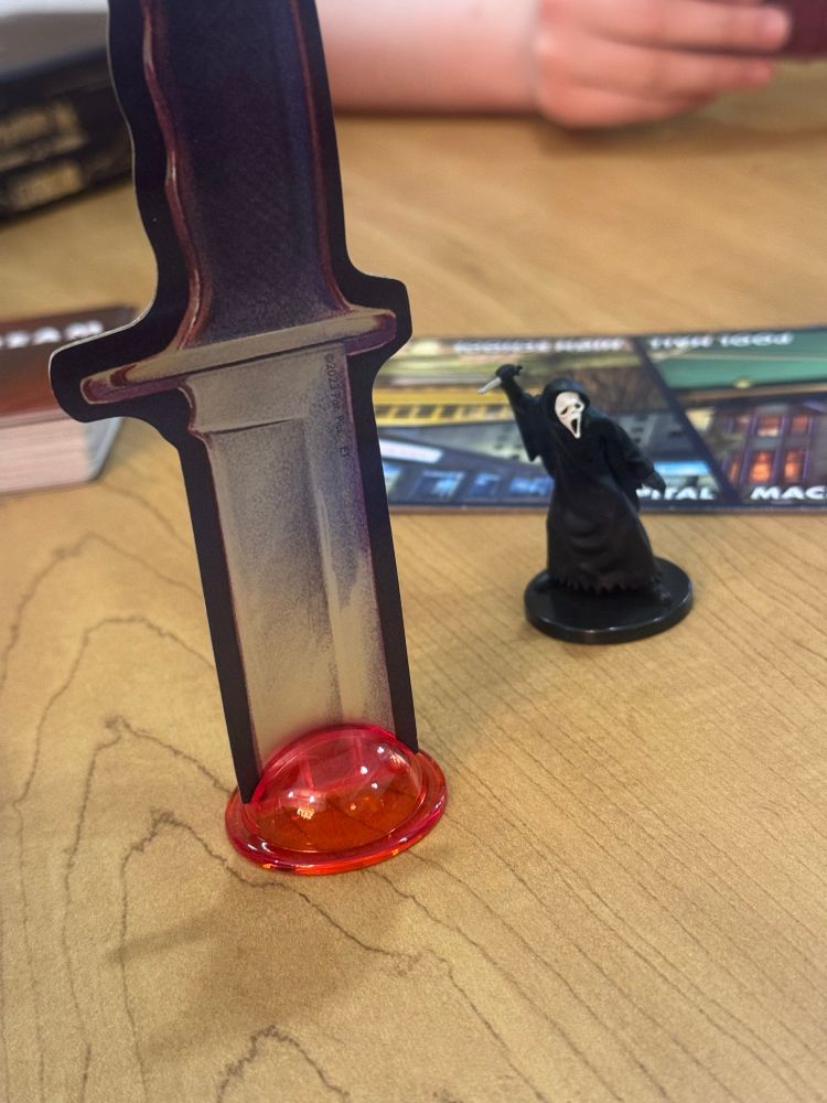 A card board knife in a translucent red holder next to a ghost face miniature from scream the game 
