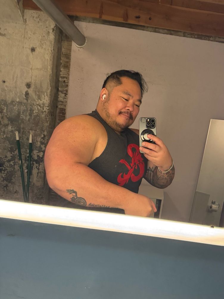 Danny, an East Asian person flexing his arm in the mirror with a slight smile wearing grey tank top with a red dungeons and dragon’s ampersand logo