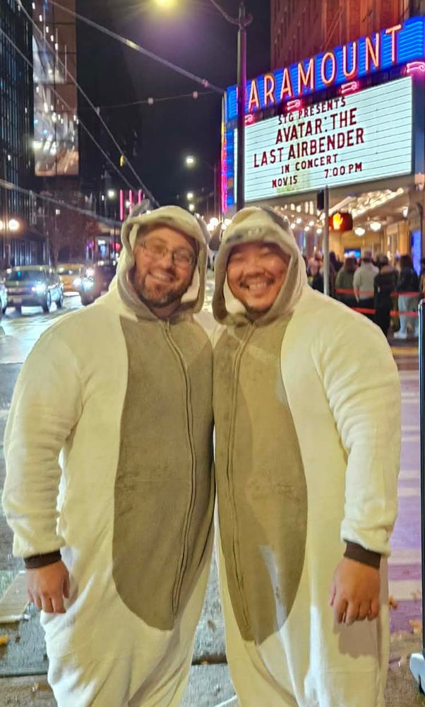 Joe and danny in appa onesies (air bison from a cartoon) in front of a theater that reads avatar the last airbender in concert 