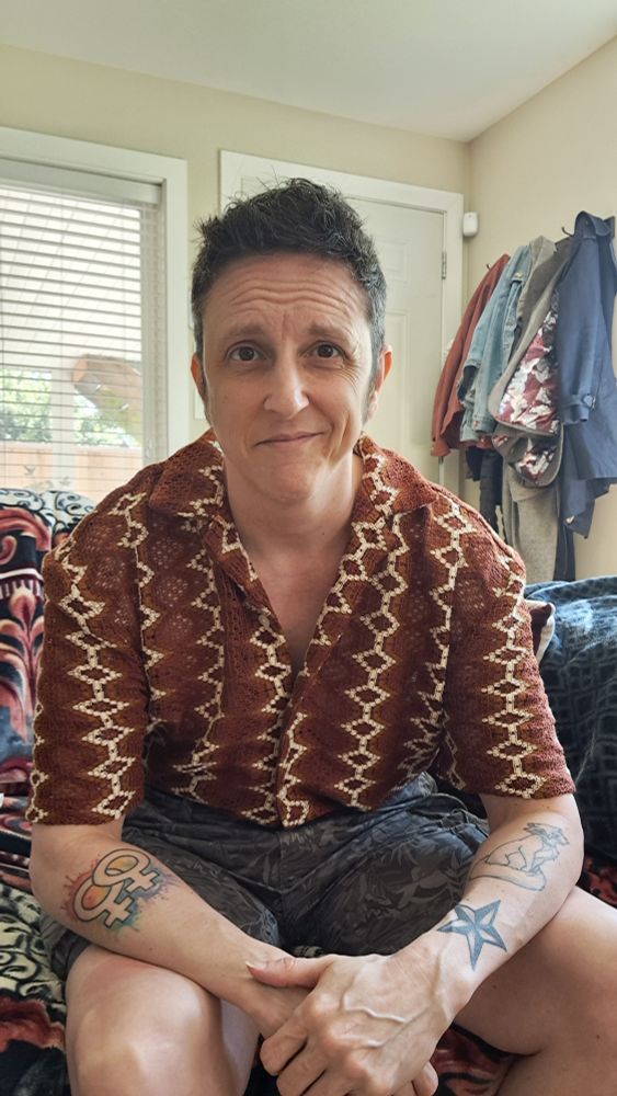 A white guy with short brown hair, brown eyes, and tattoos on his forearms, wearing a muted orange and white open knit shirt and grey cargo shorts, sits on a couch smiling.