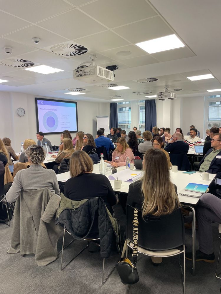 A room full of people seated at tables during TASO's 'Developing a whole-provider approach' event in Leeds. A presentation slide titled “WPA: Previous research” is projected at the front, and attendees are listening and taking notes.