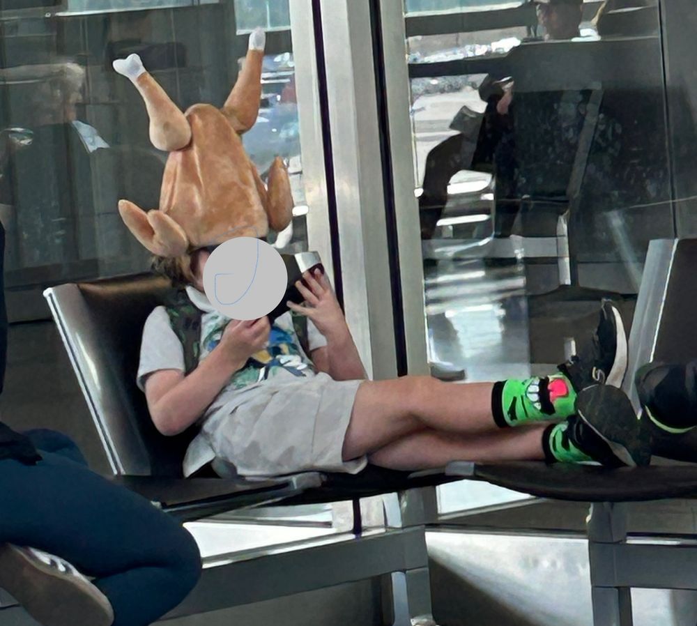 A young person lounges on a chair in an airport waiting area, their feet on a chair nearby. The person is wearing short pants, and the black sneakers are complemented by socks that are a green monster face with a bright pink tongue. The outfit is topped off with a “hat” that appears to be a large roasted turkey inverted on the child’s head. The turkey is roughly the size of the person’s trunk. 