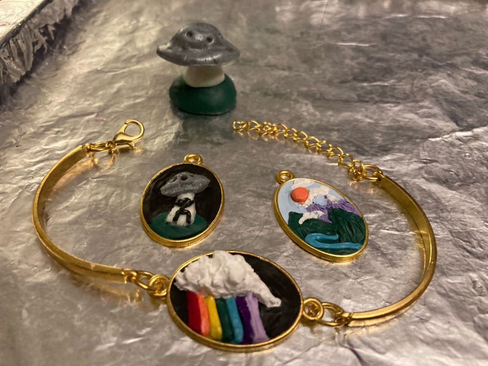 Photo of a tin baking sheet with 3 gold oval pendants with polymer clay reliefs inside. One is of a UFO abducting a stick figure man, one is of a mountain landscape, and the lady is of a textured 3D cloud on a black background with a rainbow streaming from it.
Behind the pendants is a sculpture of a silver UFO floating above a green hill.