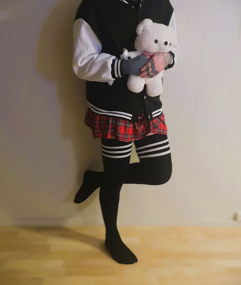 Femboy holding a white teddy bear backpack, wearing a varsity jacket, red checkered skirt and black thigh highs with three white strupes around the thigh