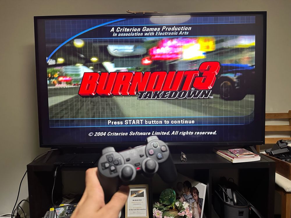 The title screen of “Burnout 3: Takedown” for PS2 on a large flat screen TV, with my hand holding up a PS2 controller in front of it. 