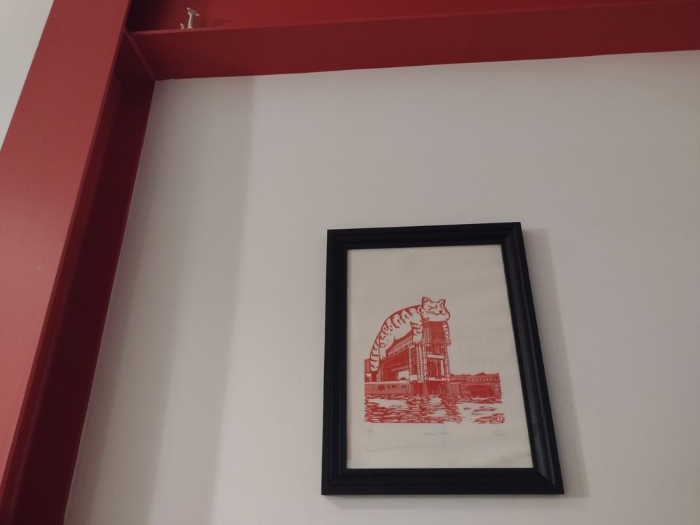 estampe d'un gros chat qui dort sur le ministère des finances (à Bercy), imprimée en rouge dans un cadre noir, sur un mur avec deux poutrelles métalliques peintes en rouge elles aussi 