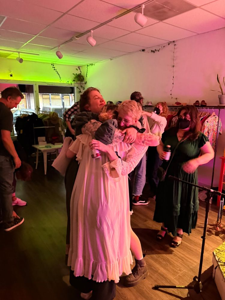 the three of us hugging after the show. the tiny venue is a thrift store by day, so you can see the racks of clothes in the background. it's mostly empty after the show, but there's a couple people still hanging around. we all look overjoyed. i am holding a can of seltzer.