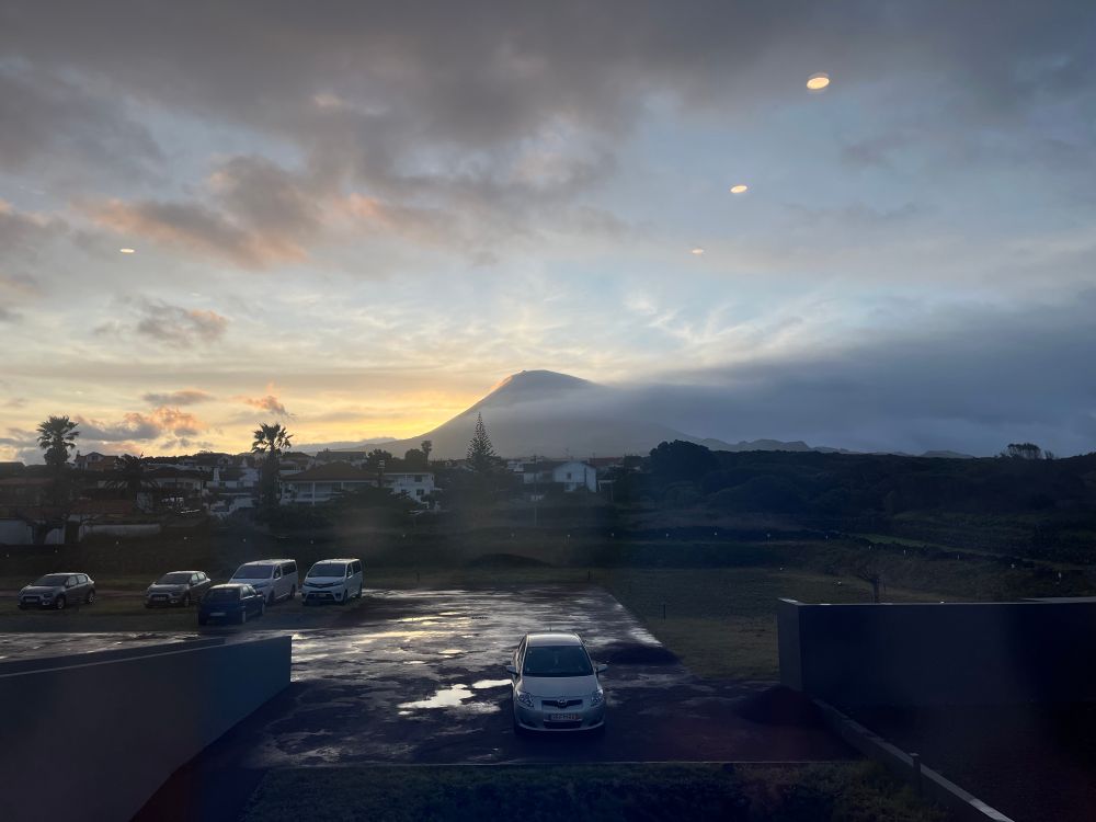 Sunrise view from a building, with a parking lot in the foreground, palm trees and other foliage mixed with residential houses. In the background, Mount Pico has a blanket of cloud trailing down the far side while the sun rises behind the left slope 