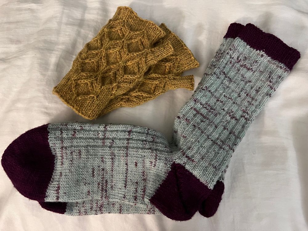 A pair of fingerless gloves in the color of honey have a honeycomb stitch pattern. They are lying next to a pair of light blue ribbed socks with dark purple toes, heels, and cuffs. Both are lying on a white blanket as the background.
