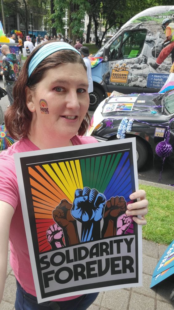 Morgan, a middle aged woman with brown hair, stands in front of several union decorated vehicles at the pride parade. She holds a sign that has a number of raised fists against a rainbow colored sunburst with the text Solidarity Forever.