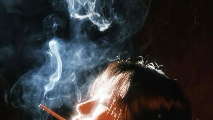 in a dark background a side profile illuminated by harsh orange light smokes a cigarette. the image is cropped just below the person's nose