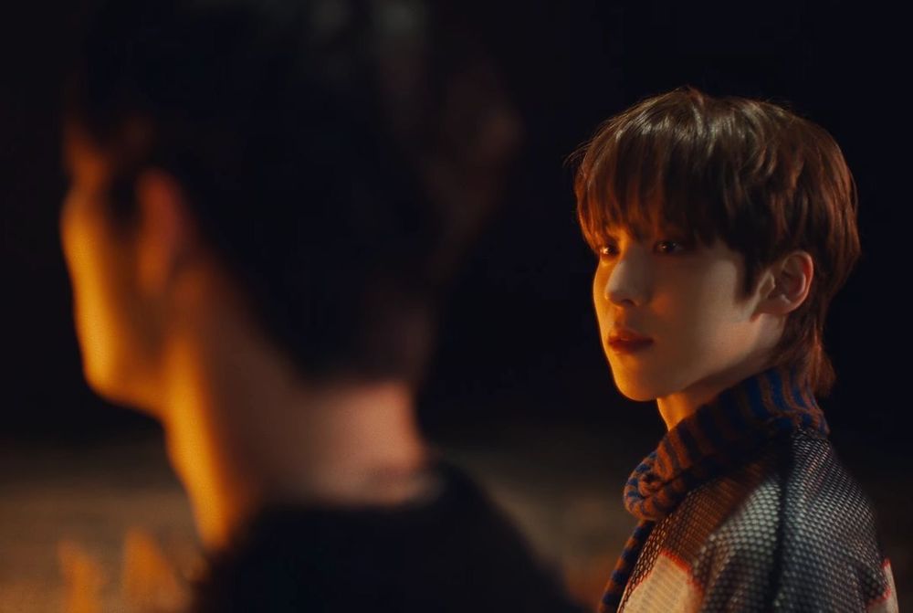 picture of yunho and mingi of ateez in low, warm lighting. yunho looks at mingi, who is out of focus, turned away from the viewer. they both seem to be facing a fire out of view.