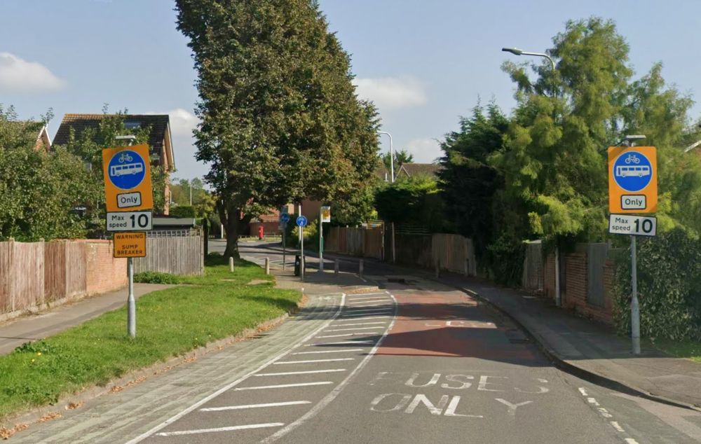 A single lane road with a cycle lane heading towards a bus stop and a sump buster, a petruding metal and concrete lump, in the background. A pair of blue and yellow pedal cycle and bus only signs are in the foreground either side of the road. Below them are 'max speed 10' signs, and a yellow warning sign for the sump buster.