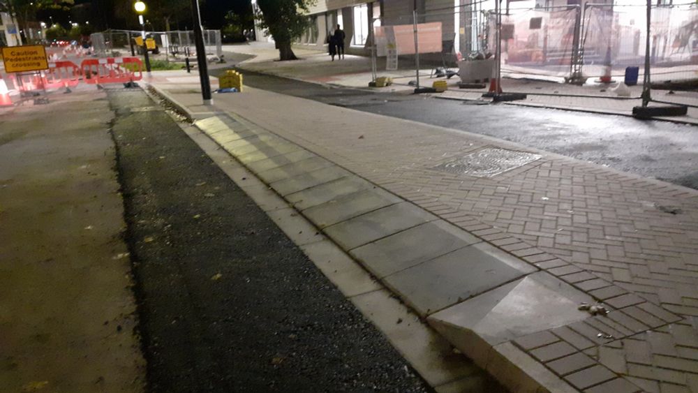 A newly constructed pavement in amongst roadworks. A number of large sloped kerbstones form a ramp along the kerbline for around 3m. Behind thr pavement is a cycleway still under construction.