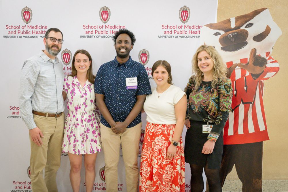 Alt text:
Five people stand smiling in front of a University of Wisconsin–Madison School of Medicine and Public Health backdrop. From left to right: a man in glasses and a checked shirt, a woman in a floral dress, a smiling man with a name tag, a woman in a white top and red floral skirt, and a woman in a black dress with colorful embroidery. To the far right is a life-size cutout of Bucky Badger, the university mascot.