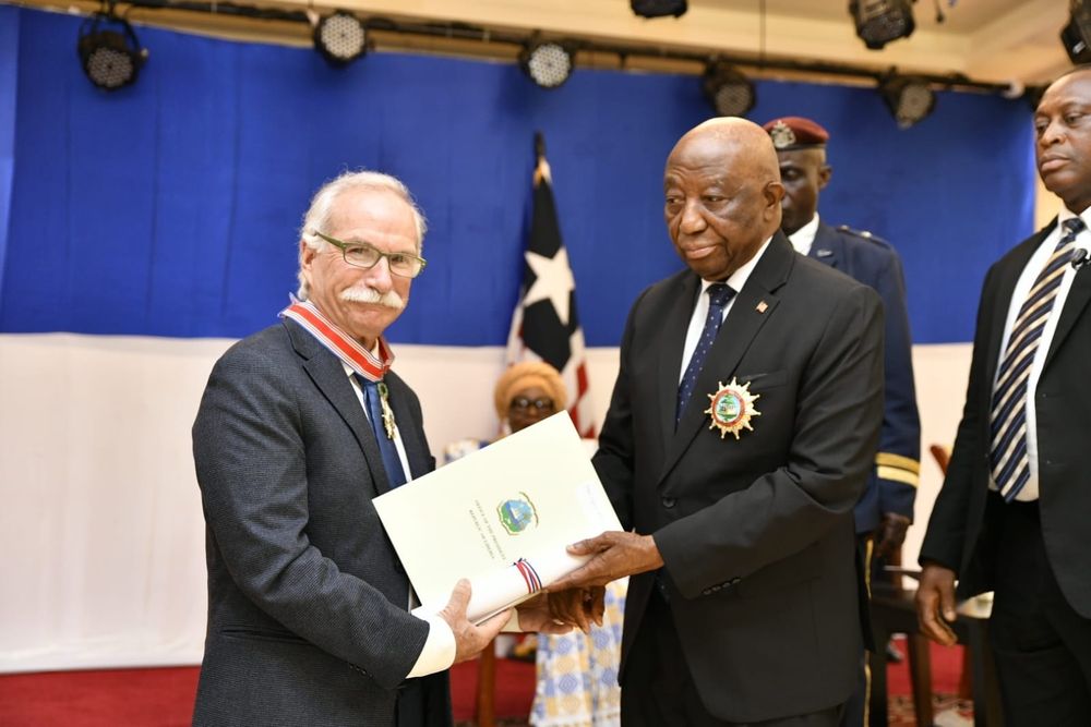 Gregg Mitman receives the Humane Order of African Redemption from Liberian Vice President Jeremiah Koung during an official ceremony. Both men wear formal attire and ceremonial medals. Mitman holds a certificate, and the Liberian flag is visible in the background.