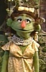 A green Muppet with glasses, curly hair, a pearl necklace, and a golden dress.