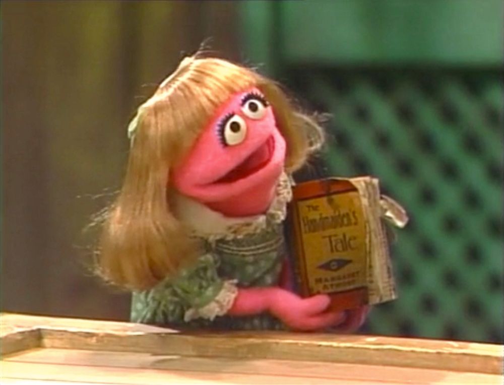 Prairie Dawn, a tiny hot pink Muppet with blonde hair and a blue dress, holding The Handmaid's Tale by Margaret Atwood.