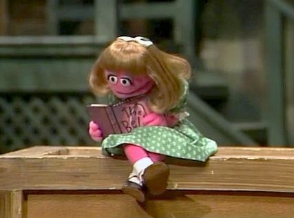 Prairie Dawn, a tiny hot pink Muppet with blonde hair and a blue dress, reading The Bell Jar.