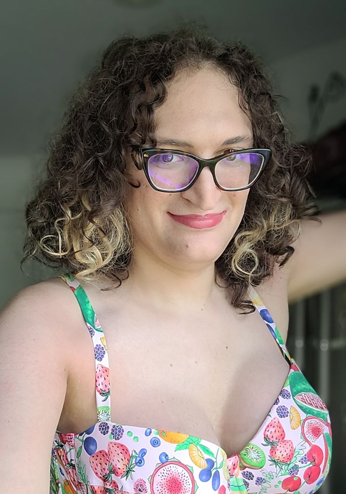 A current photo of Becca. She has long curly hair and is smiling in a fruit print top.