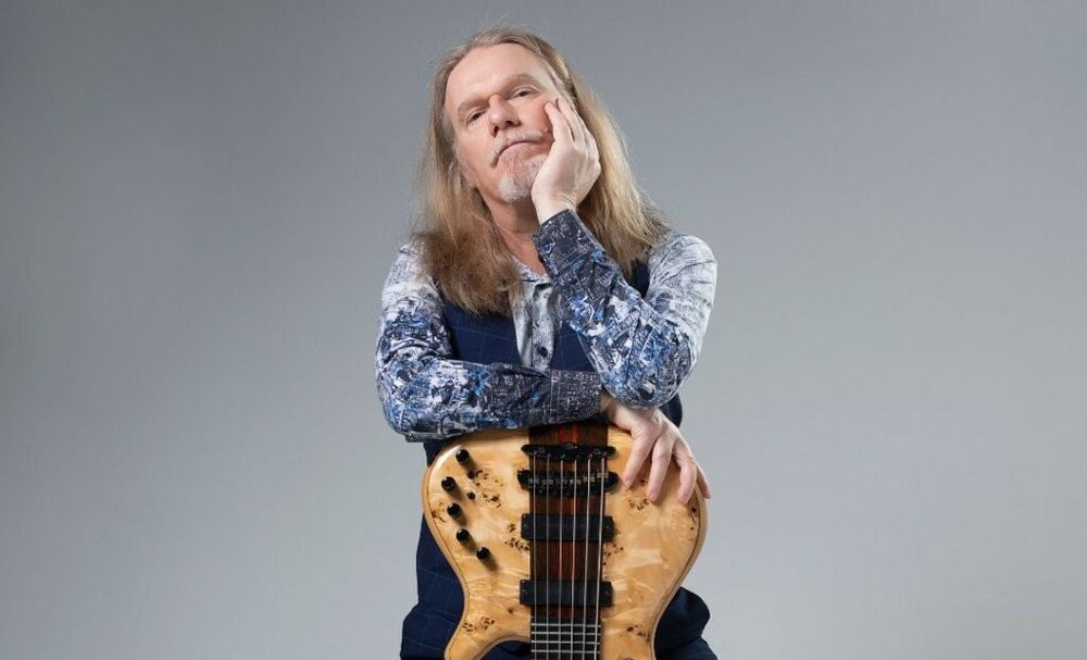 Humberto Gessinger com a cabeça apoiada na mão e cara de pensamento, seus braços estão apoiados em uma guitarra de ponta cabeça. Ele veste uma blusa estampada de manga comprida, em tons de azul e um pouco de branco. A guitarra é bege com o meio marrom e tem 6 cordas. 