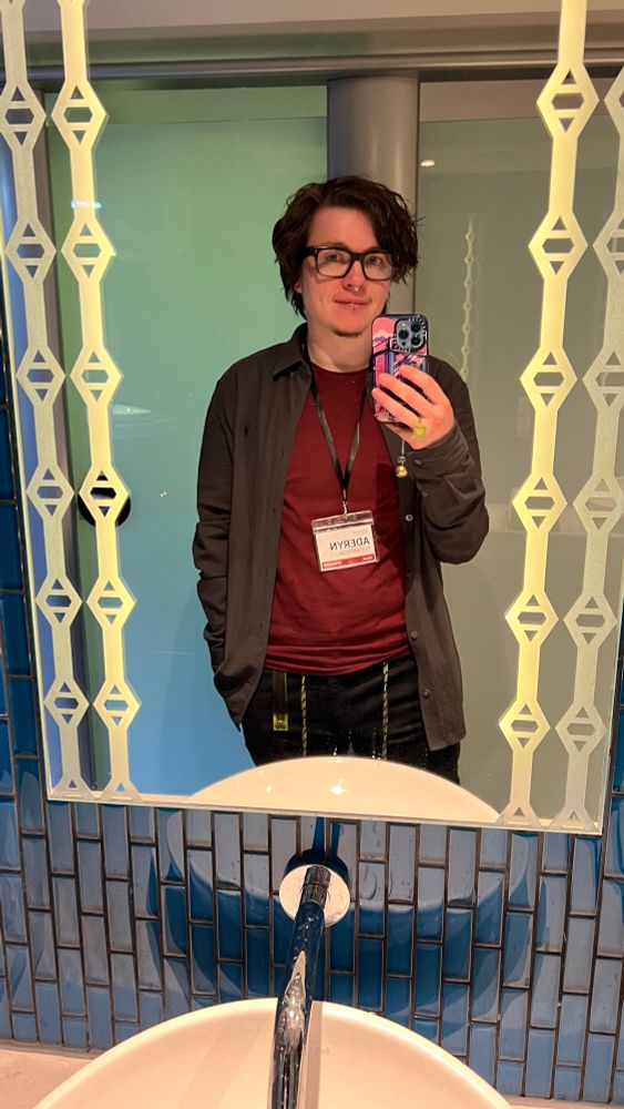 Aderyn in a mirror wearing a red shirt, grey over shirt, dark blue trousers with short curly black hair and thick dark glasses 