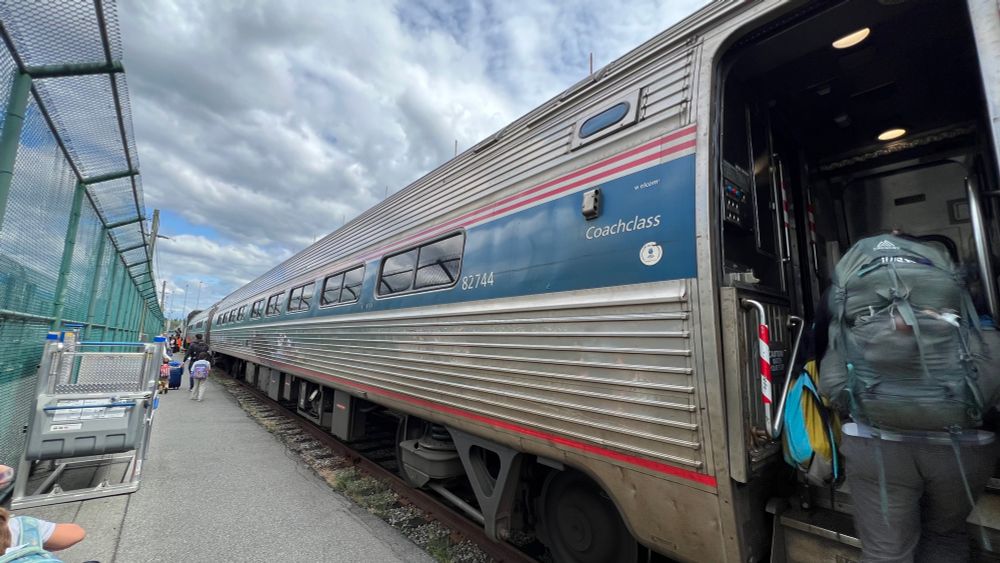 An Amtrak Cascades train car in the station. It’s silver and blue and there’s a cloudy sky above with someone climbing aboard