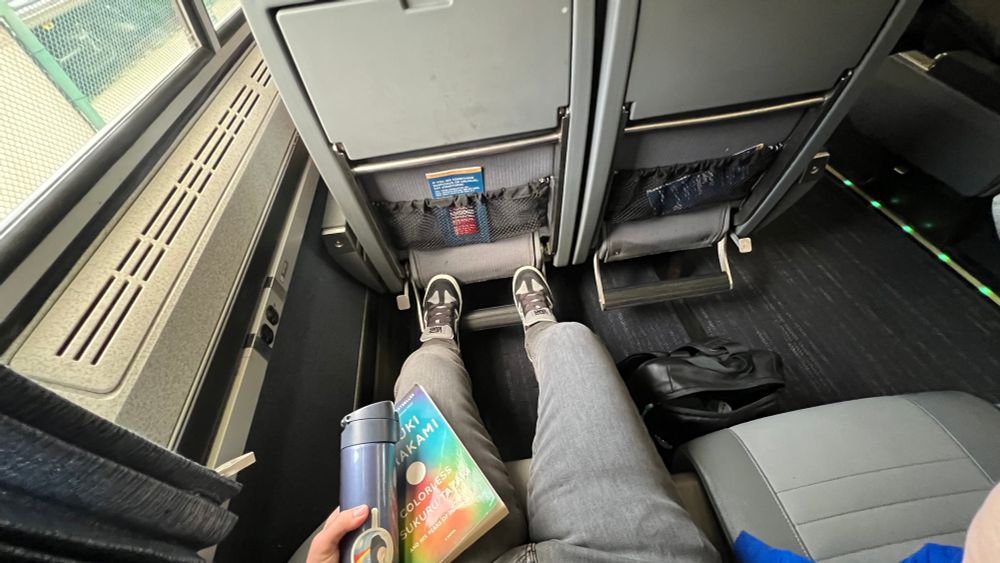 Legs in grey skinny jeans sat in a train car seat with a hand holding a Haruki Murakami book and a zojirushi travel mug 