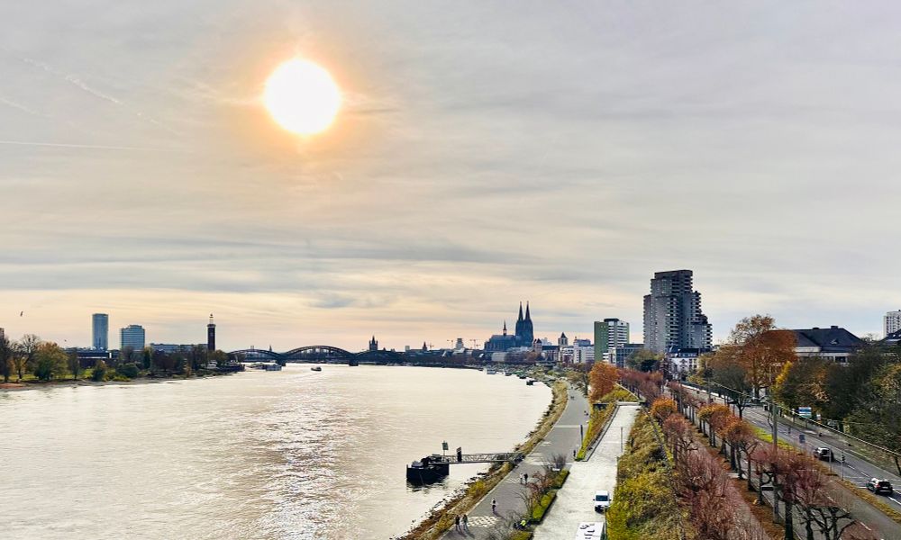 A wide Rhine River flows through Cologne, Germany under a bright sun. The iconic twin spires of Cologne Cathedral dominate the skyline. Modern buildings line both banks, with a pedestrian path along the waterfront and autumn trees adding color.
