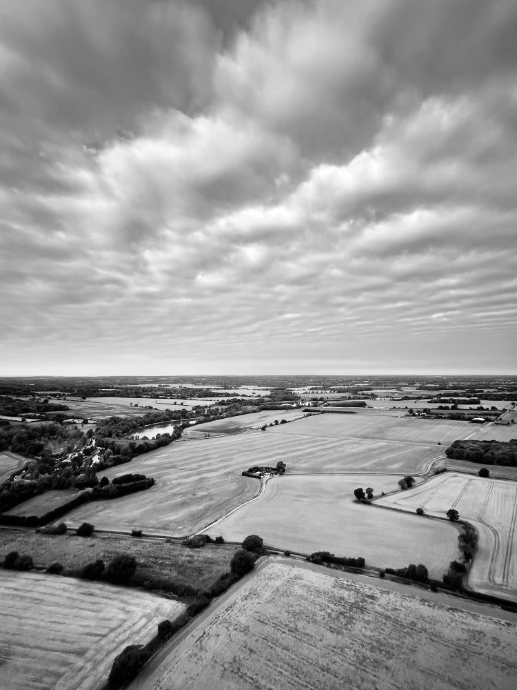 A black and white photo from a hot air balloon looking down on a patchwork of fields and hedges that seems to be split in half with the top half showing some cotton wool pleated clouds stretching out above the horizon.