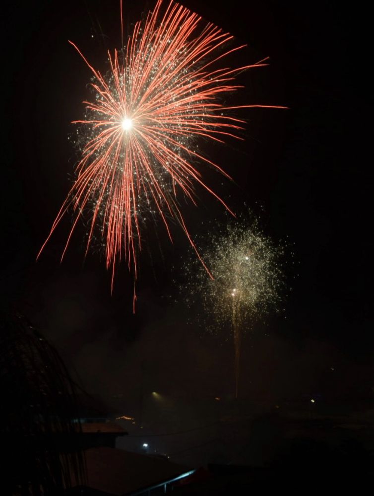 Midnight on New Year’s Day 2025 in Kaneohe, HI. A dimly lit, smoky neighborhood with the ghostly silhouette of a house in the lower left corner. In the upper left corner a firework explodes, looking reminiscent of the pinkish ribbons that wrap around Sailor Moon when she transforms. A second, sparklier, white/yellow firework explodes in the background of the first, in the right-ish center of the frame. The rest of the photo is black. 