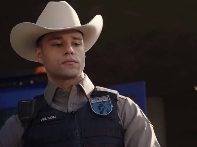 Carlos Reyes, de 911 Lone Star, vestido como Texas Ranger, colete à prova de balas e chapéu de caubói. Ele está com um semblante sério, olhando para o suspeito.
