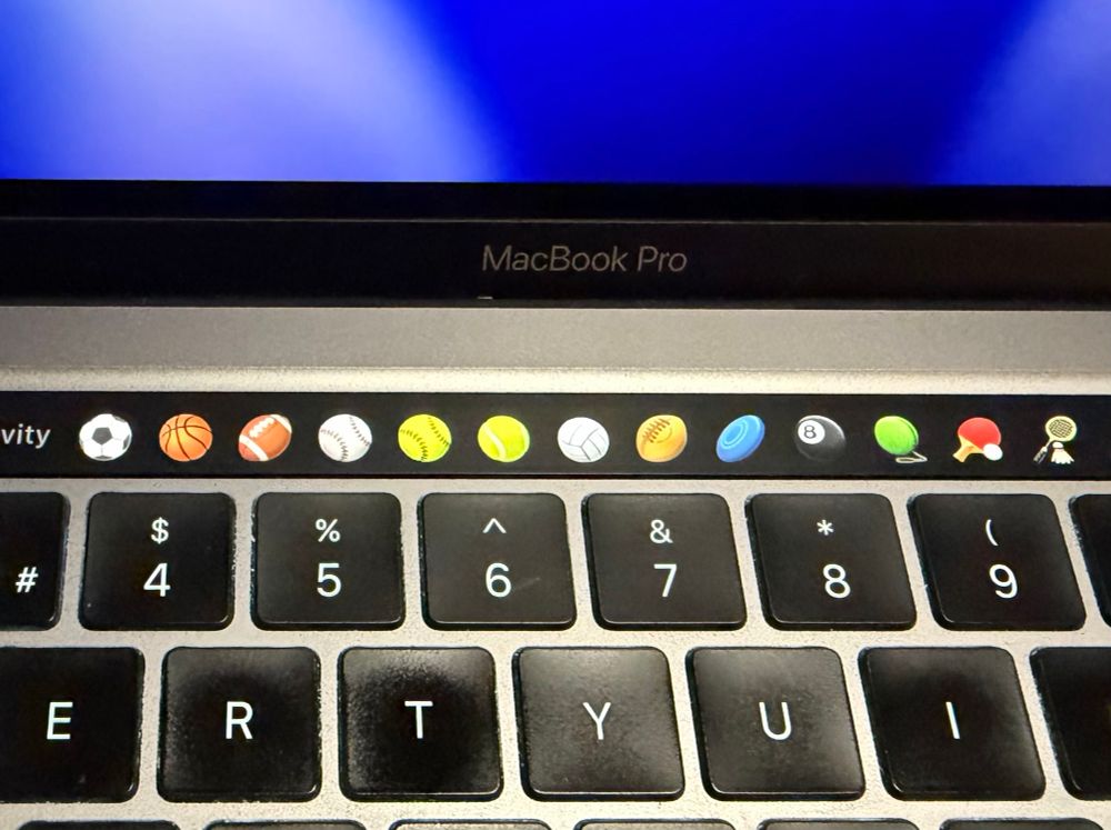 This photo shows a MacBook Pro with the Touch Bar activated and displaying a row of sports-related emojis. The screen is partially visible at the top, showing a gradient blue background. The Touch Bar, located above the number keys, features various sports ball icons including soccer, basketball, American football, baseball, softball, tennis, volleyball, rugby, flying disc (Frisbee), billiards 8-ball, and table tennis, as well as a green ball, a red ping pong paddle, and a badminton racket with shuttlecock. The MacBook Pro branding is visible above the Touch Bar.