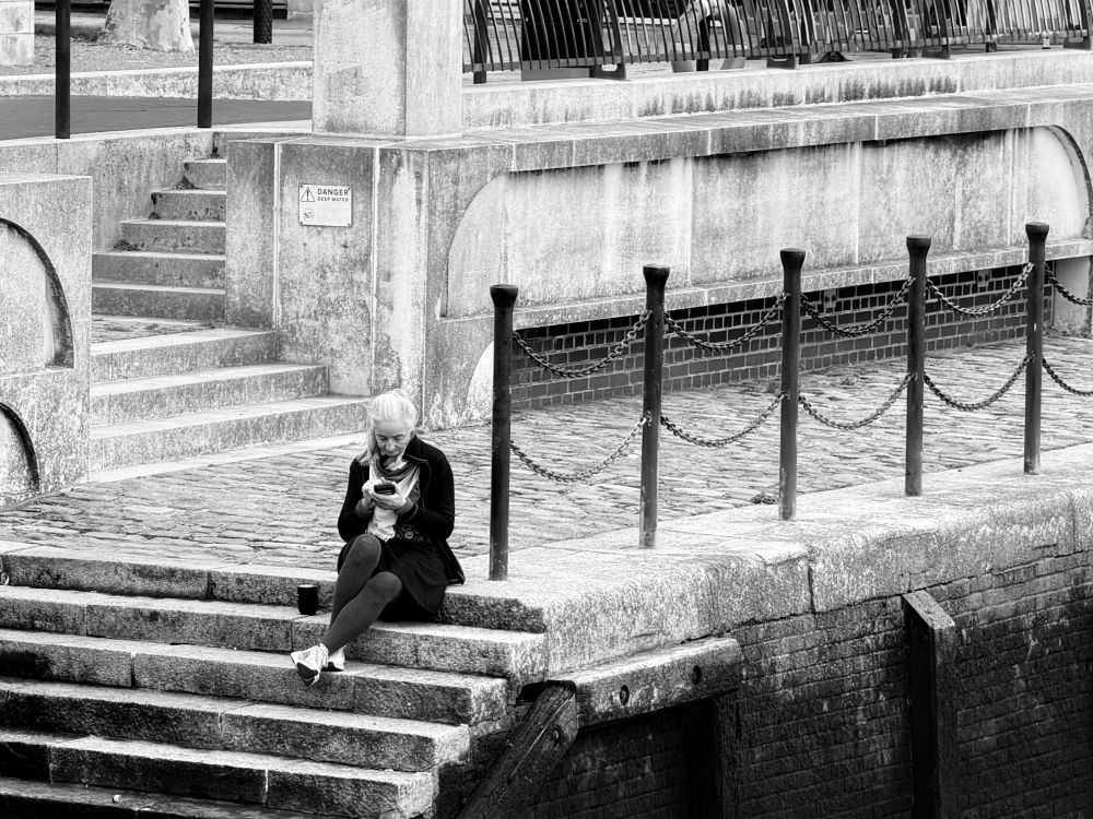 This photo is a black-and-white image showing a solitary figure sitting on stone steps beside a waterfront or canal. The individual appears relaxed, focused on a mobile device held in their hands, and has a cup or container placed beside them on the step. The background consists of rough stone walls, iron posts linked with chains, and more steps leading up to a street level above. A sign saying “DANGER” is visible on the wall behind, suggesting the area may be adjacent to deep water. The overall atmosphere is calm and slightly industrial, combining the textures of old stone, metal, and brickwork. The person’s face has been deliberately blurred to maintain privacy. 