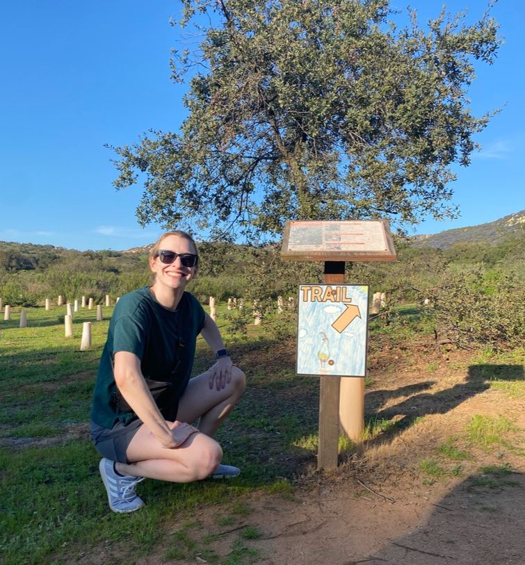 Gabi is wearing sunglasses and smiling outside, in front of a sign with a child's drawing of a quail, an arrow and a TRAIL label. 