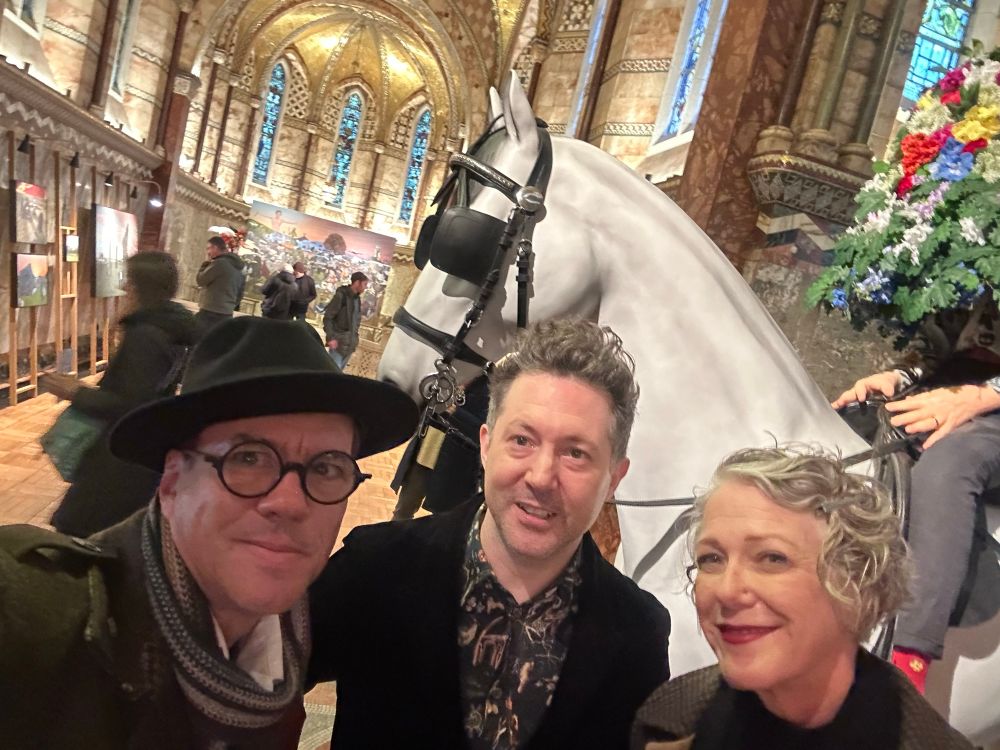 Two men and a woman posed in front of a sculpture of a white horse ridden by a man covered in flowers. 