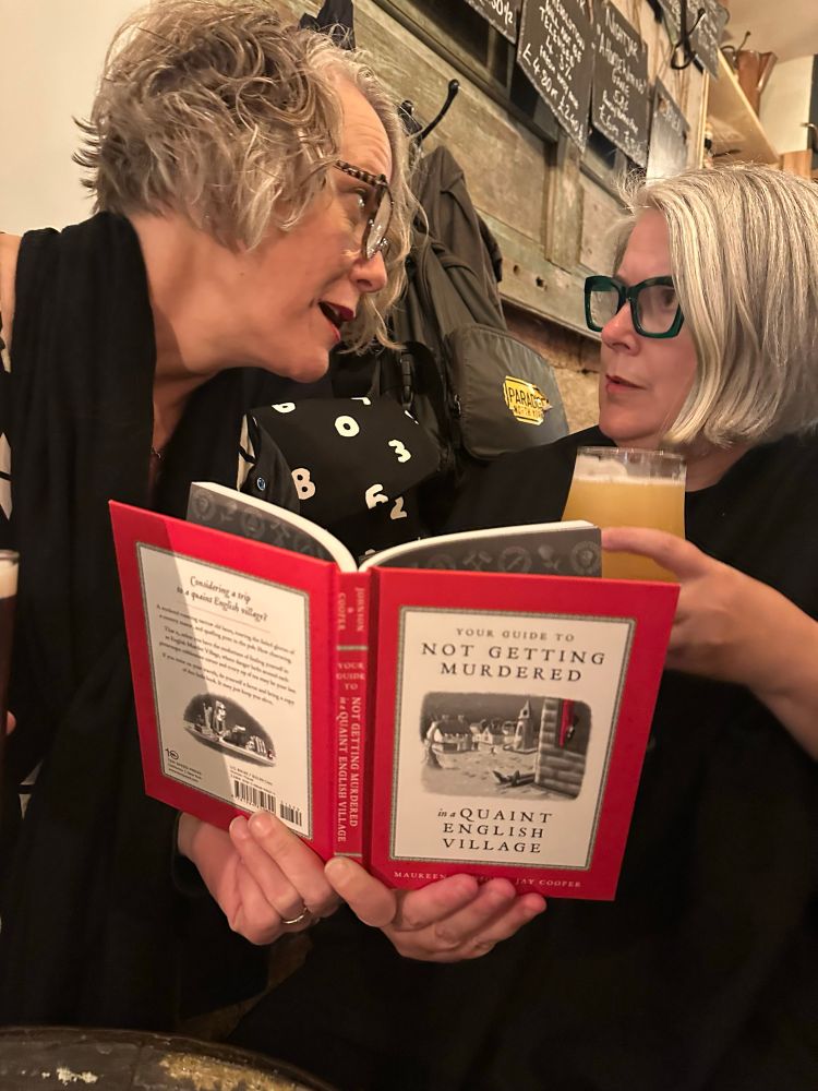 Two women in glasses reading the book ‘Your Guide to Not Getting Murder in a Quaint English Village.’