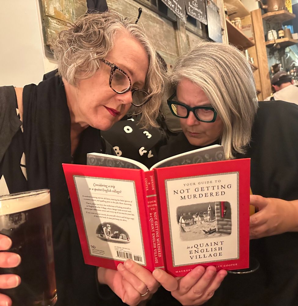 Two women in glasses reading the book ‘Your Guide to Not Getting Murder in a Quaint English Village.’