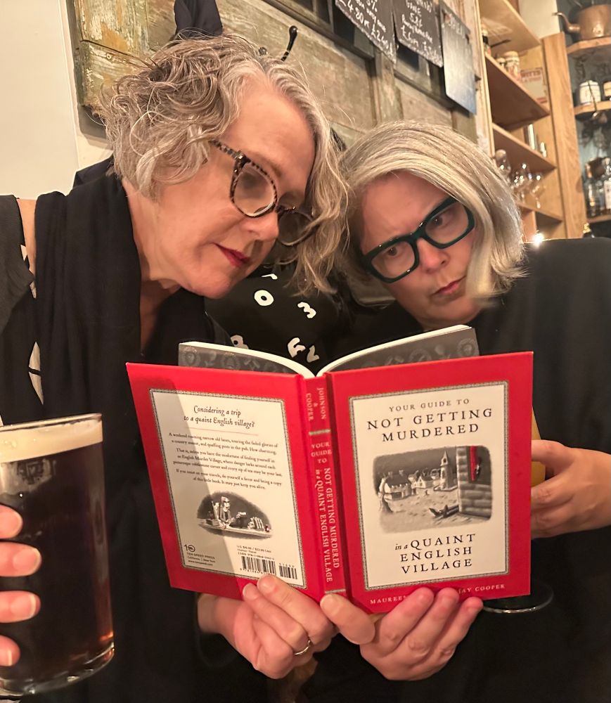 Two women in glasses reading the book ‘Your Guide to Not Getting Murder in a Quaint English Village.’