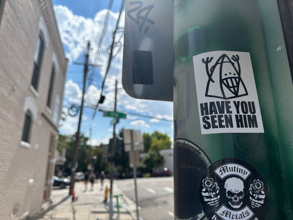 A sticker on a post with a small triangular icon with the text: Have you seen him