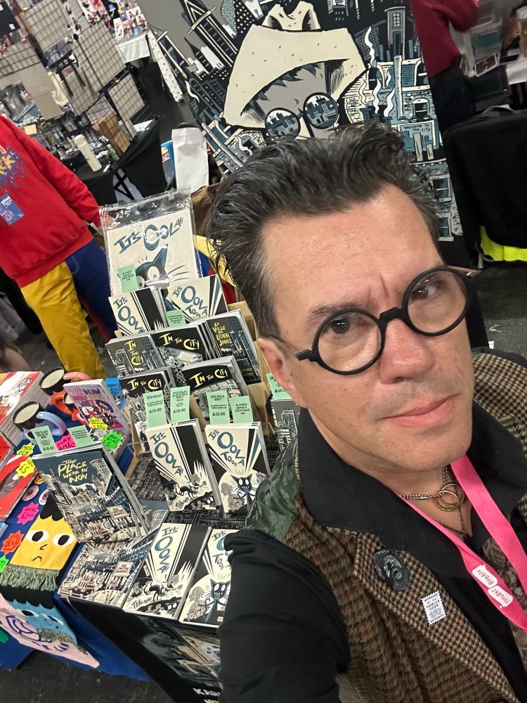 A man in glasses standing by a table filled with books and comics.