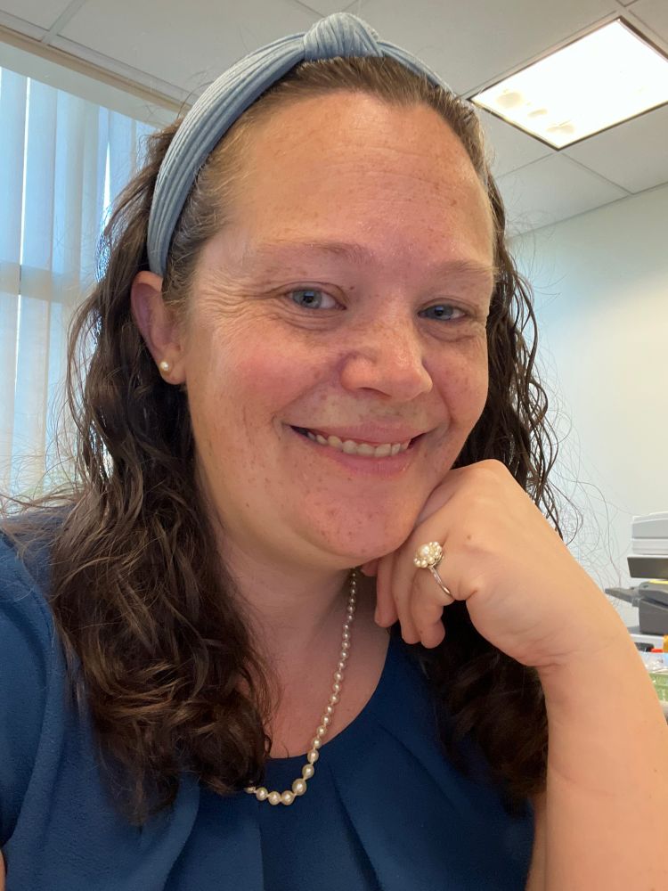 A white woman with curly brown hair and blue eyes smiles at the camera. She’s wearing a blue blouse, pearl jewelry and a light blue headband, and resting her chin on her hand to show off her pearl ring.