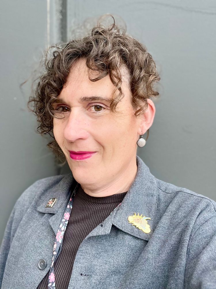 Siân, a white trans girl-ish, in a grey shirt over a dark grey dress, nhw hair is curling madly